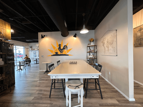 A spacious, modern café interior with a large table, black chairs, and a rooster mural on the wall.